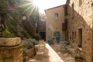 A courtyard with stone flooring, metal chairs, and tables, surrounded by stone walls and vegetation.