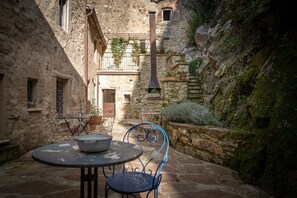 A stone courtyard with a table and chairs, a rustic building with a chimney, and a narrow passageway.