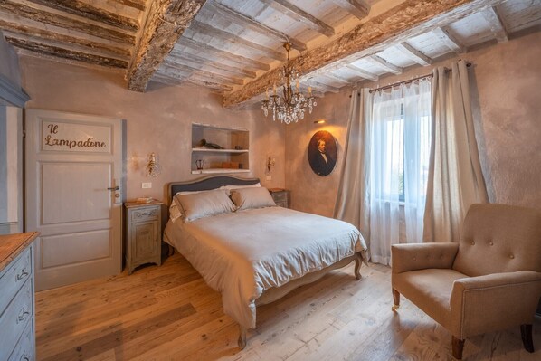 A bedroom with a bed, a chair, a nightstand, a dresser, a chandelier, a portrait, and a door labeled 'Il Lampadone.'