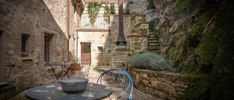 Une cour pavée de pierres avec une table et des chaises, un bâtiment rustique doté d’une cheminée et un étroit passage.