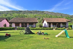Eine Rasenfläche mit Spielgeräten, zwei Häuser mit Ziegeldächern und eine hügelige Landschaft im Hintergrund.