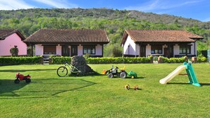 Un espacio con césped, equipo de juegos, dos casas con tejados de tejas y un paisaje montañoso al fondo.