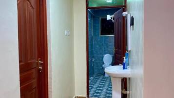 Un pasillo estrecho con una puerta de madera a la izquierda, un lavamanos blanco con espejo a la derecha, y una zona de ducha con azulejos azules al fondo.