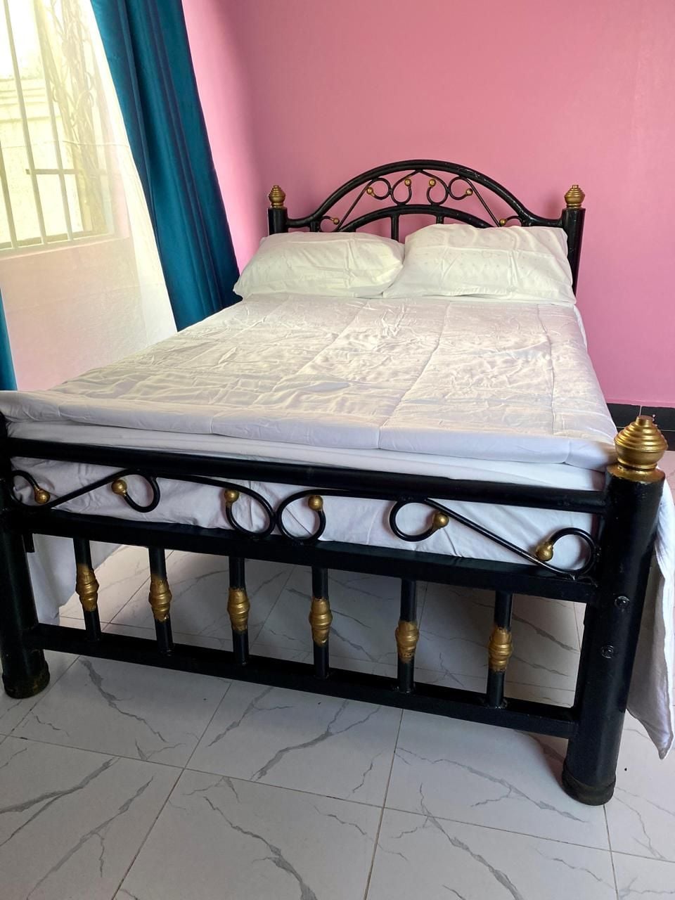 A bed with a black metal frame and gold accents, set against a pink wall.