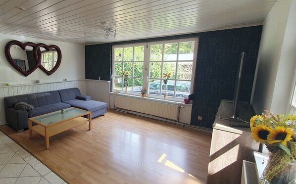 A living room with a sofa, coffee table, and a large window with a view of parked cars and greenery outside. - Herzkate Vacation Rental (Bremervörde)