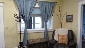 Una habitación con ventana, un sillón de cuero negro, un baúl antiguo y una lámpara de pie.