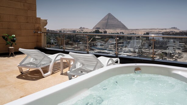 Een dakterras met een jacuzzi, witte lounge fauteuils en uitzicht op de piramide.