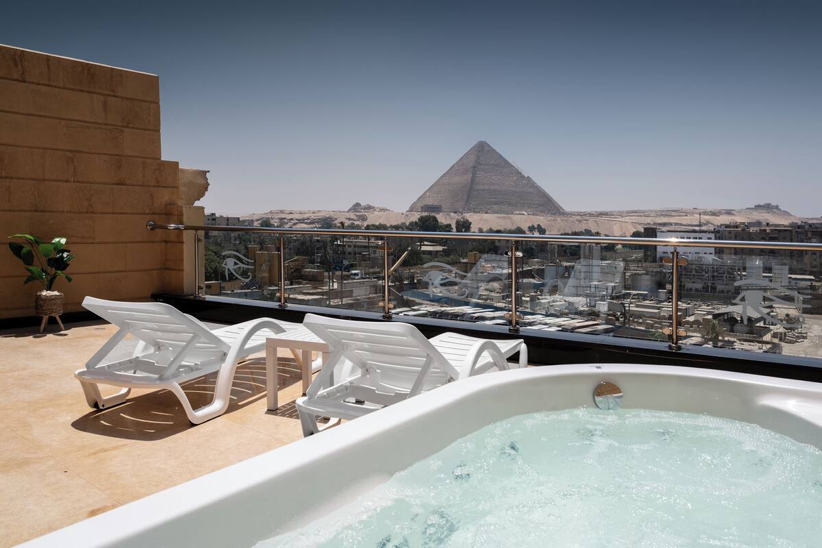 A rooftop terrace with a hot tub, white lounge chairs, and a view of the pyramid.