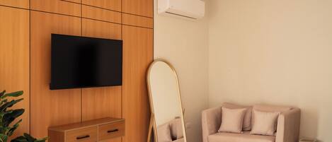 Une chambre moderne avec un téléviseur à écran plat fixé sur un mur en panneaux de bois, un petit meuble en bois avec des tiroirs, un canapé beige, un grand miroir et un appareil de climatisation blanc.