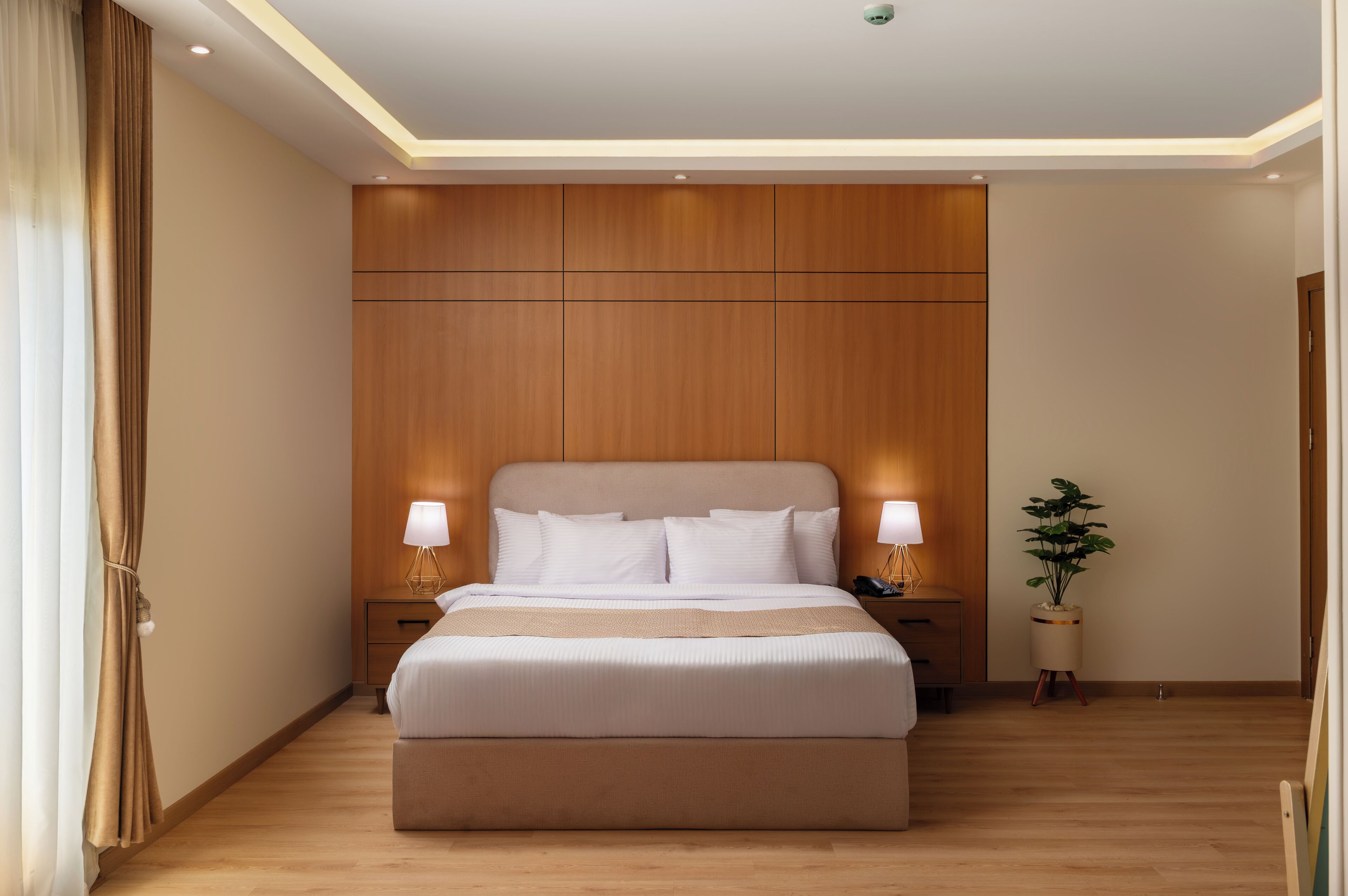 Quarto de hotel moderno com uma cama grande, cabeceira de madeira, duas mesas de cabeceira com abajures, um criado-mudo e um vaso com planta.