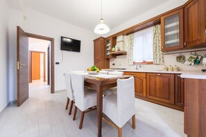 A modern kitchen with wooden cabinets, a dining table with chairs, a TV mounted on the wall, and a window with curtains.