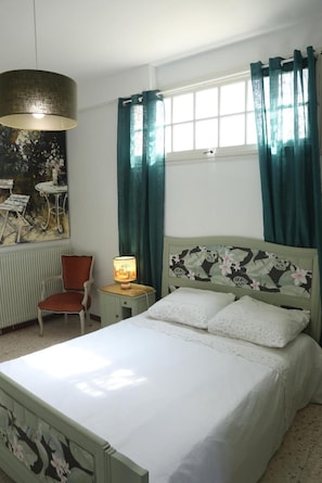 Une pièce comprenant un lit, une chaise, une table avec une lampe et une fenêtre avec des rideaux.