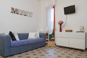 A living room with a sofa, a TV mounted on the wall, a cabinet, and a patterned floor. - Holiday Home 'Cala Lupo' with Private Terrace, Private Garden and Air Conditioning (Stintino)