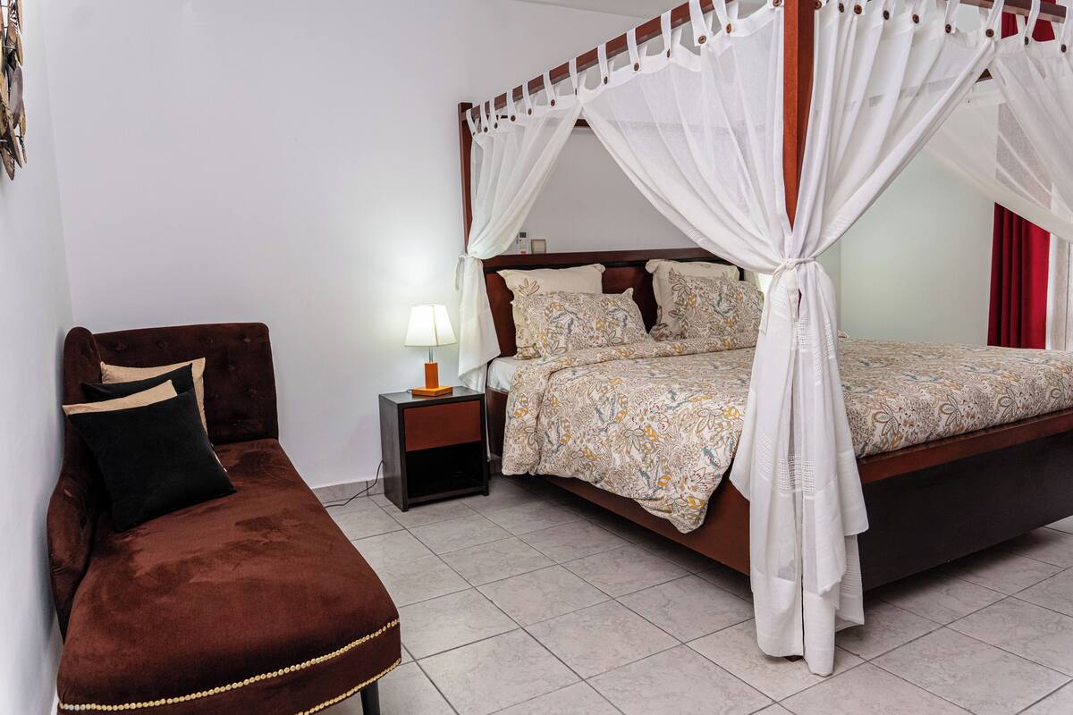 A bedroom with a four-poster bed, a nightstand, a brown chair, and a bedside table with a lamp.