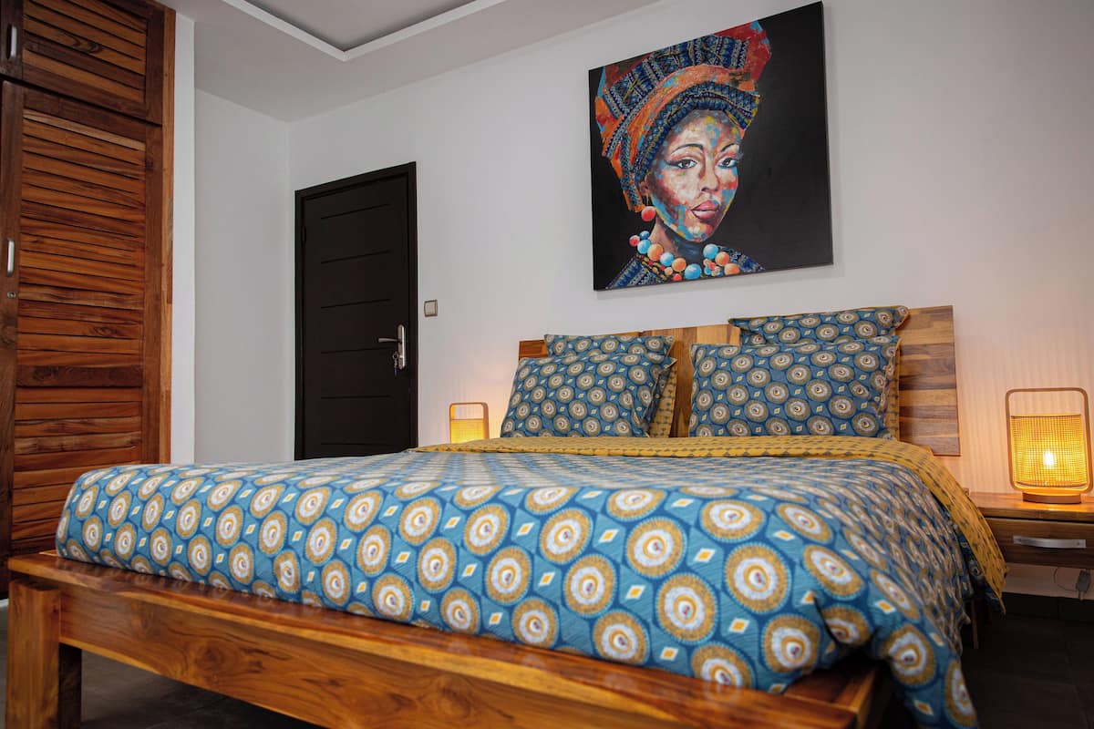 A bedroom with a bed, wooden headboard, patterned bedding, a painting on the wall, and a bedside lamp.