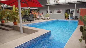 Una piscina rectangular con mosaicos azules, rodeada por un patio empedrado y un paraguas rojo. Hay sillones de playa y una mesa con sillas en una zona sombreada.