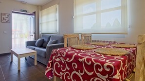 Une pièce comprenant un canapé, une petite table, une table à manger recouverte d’une nappe à motifs rouges, et une fenêtre donnant sur les montagnes.
