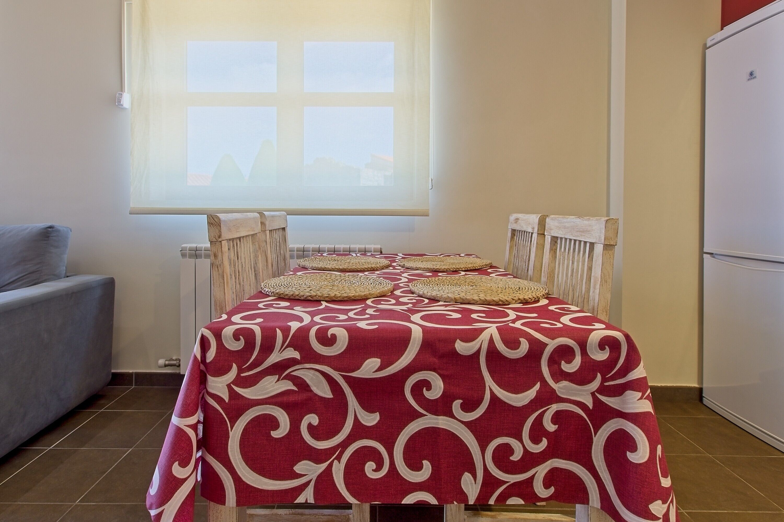 Une table à manger recouverte d’une nappe à motifs rouges et blancs, quatre sets de table en tissu, et une fenêtre donnant sur des immeubles.