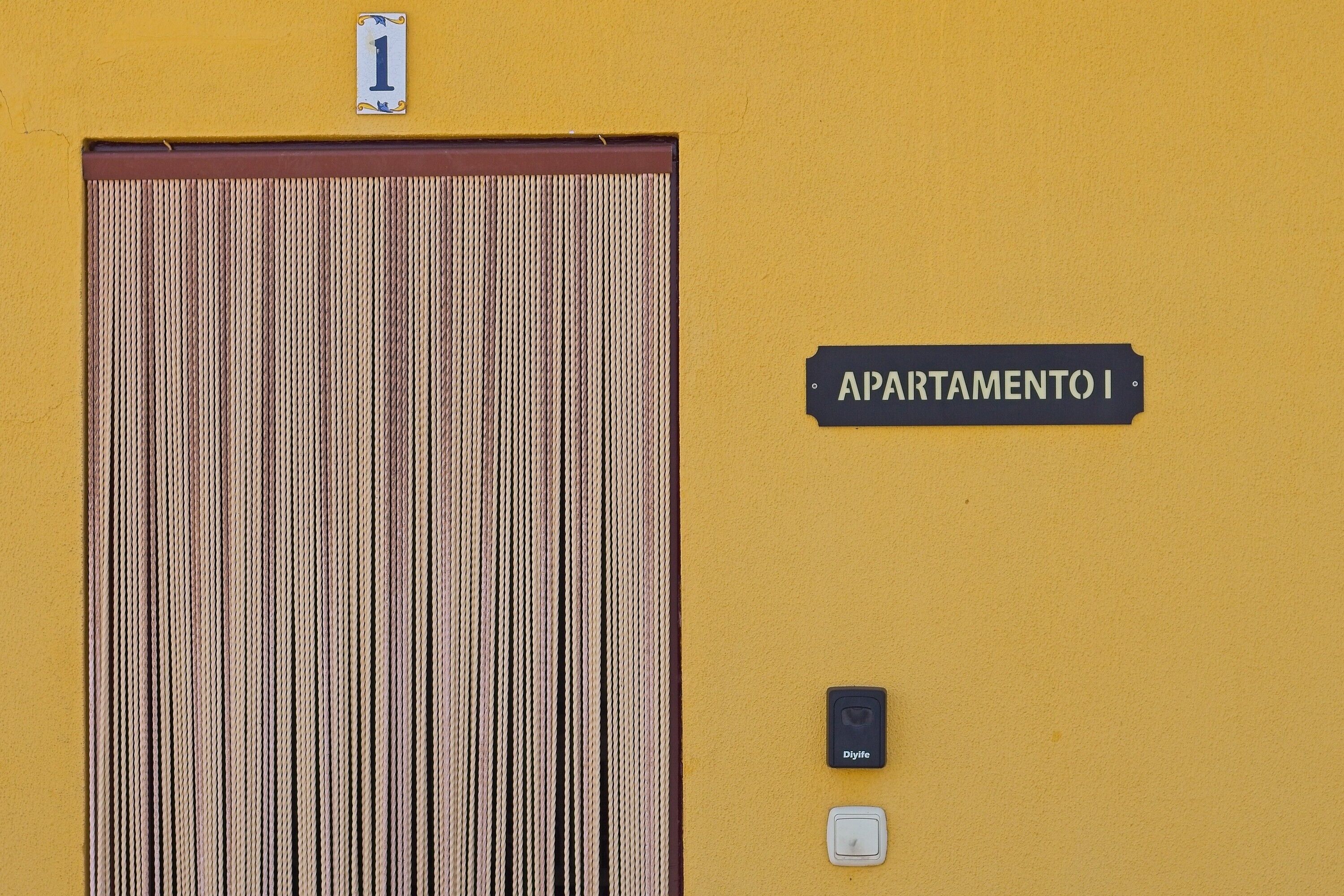 Una pared amarilla con una puerta a rayas, un timbre, un interruptor de luz y un letrero que dice 'APARTAMENTO I'.