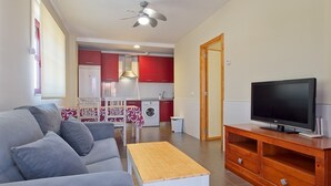 Un salon avec un canapé, un téléviseur posé sur un meuble en bois, une cuisine à l’arrière-plan et un ventilateur de plafond.