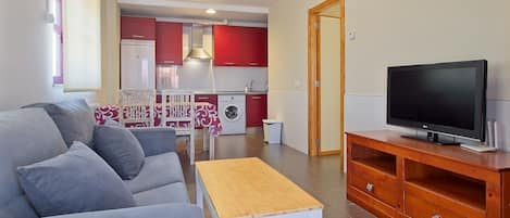 Un salon avec un canapé, un téléviseur posé sur un meuble en bois, une cuisine à l’arrière-plan et un ventilateur de plafond.