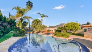 Une piscine aux carreaux bleu clair, entourée de palmiers et de verdure, avec des montagnes en arrière-plan.