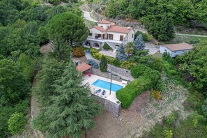 Une grande maison avec une piscine, entourée d’une végétation luxuriante et bordée d’un chemin en pierre.