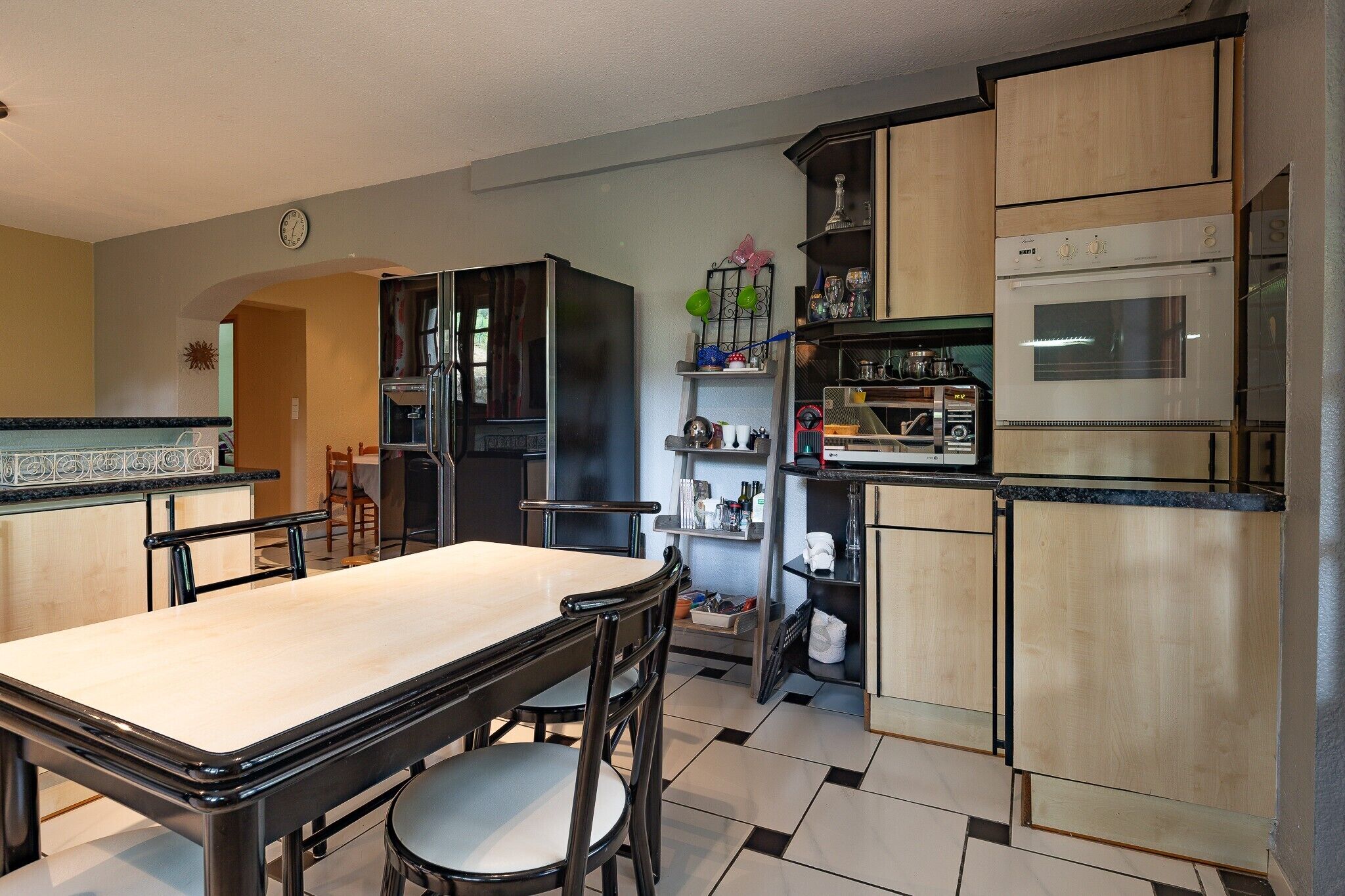 A kitchen with a dining table, chairs, a refrigerator, a stove, and shelves with various items.