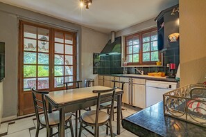 A kitchen with a dining table, chairs, a dishwasher, and a window with a view of greenery. - Apartment 'Chataigniers' with Mountain View, Private Pool and Wi-Fi (Juvinas)