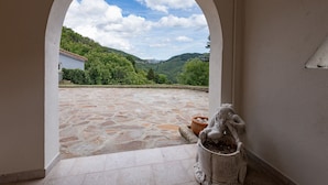 Uma porta em arco dá acesso a um pátio com chão de pedra, um vaso de planta e vista para uma encosta.