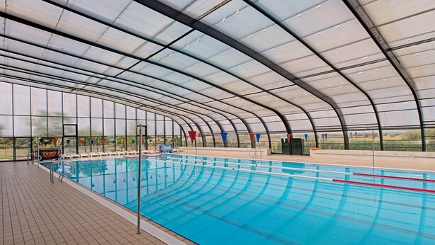 Una piscina interior con techo curvo, paredes de vidrio transparente y suelo de baldosas.