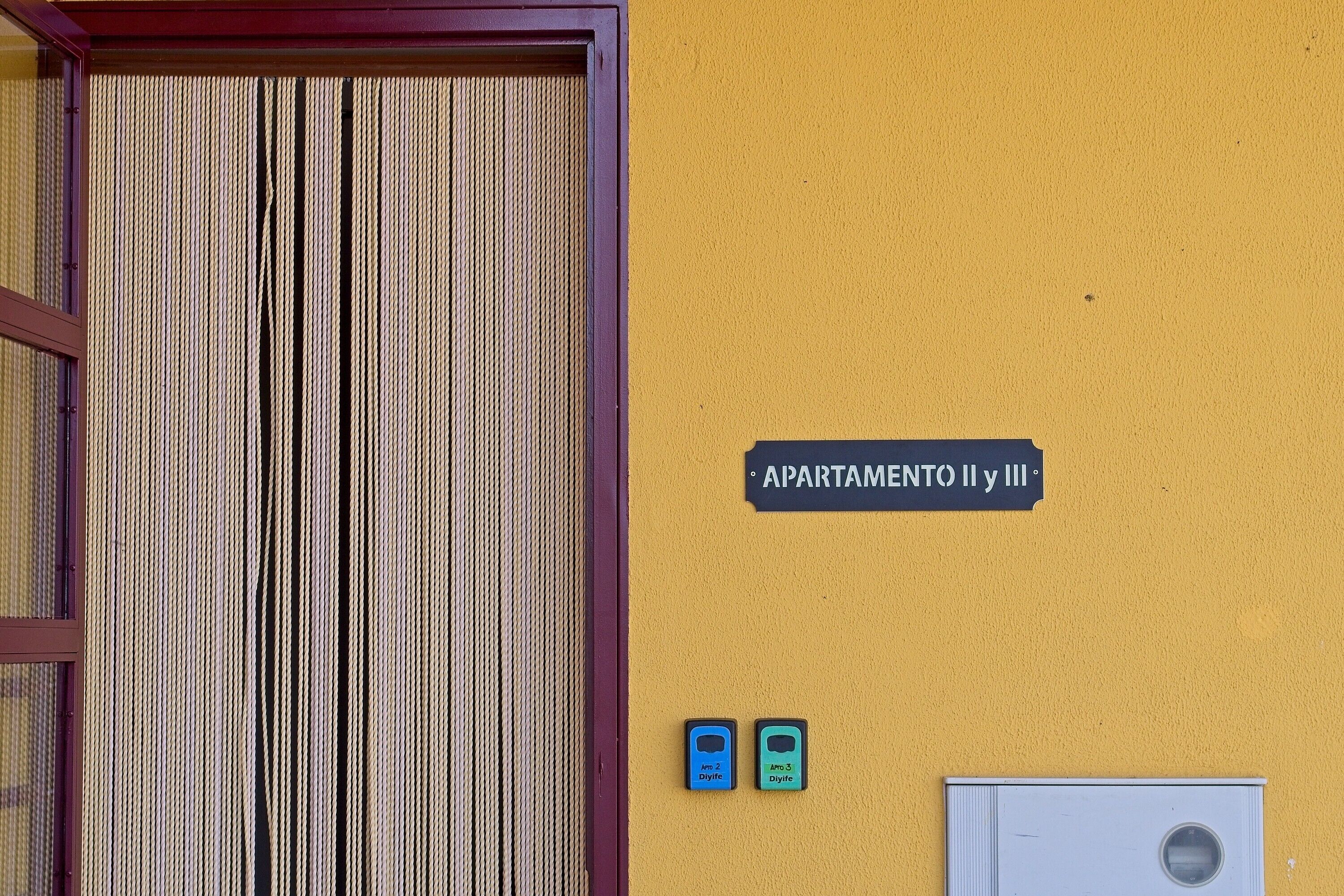 Un mur jaune sur lequel est apposée une pancarte indiquant « APARTAMENTO II y III », et deux petits appareils bleus placés en dessous.