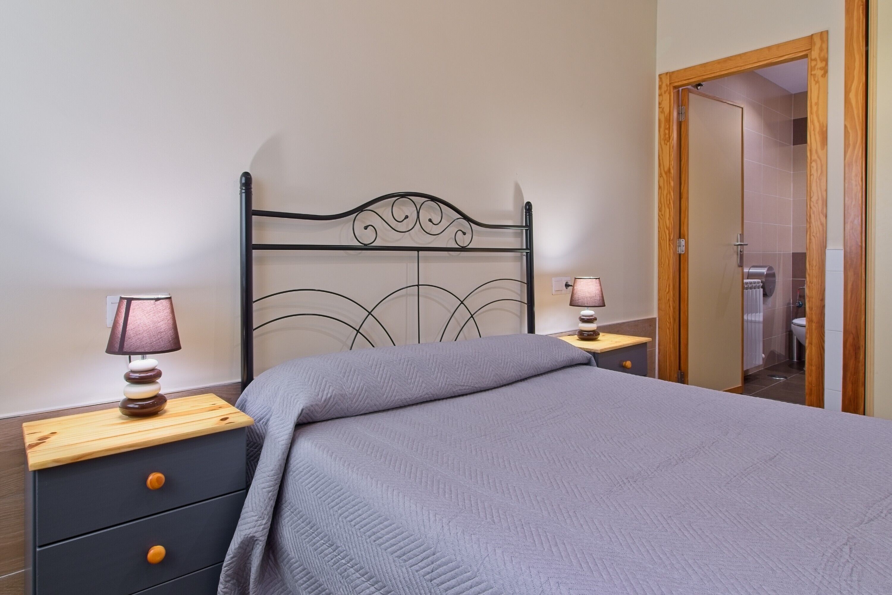 Un dormitorio con cabecera de metal, una cama con manta gris, dos mesitas de noche con lámparas y una puerta de madera que da a un baño.
