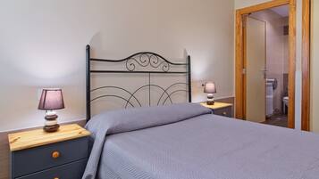 Un dormitorio con cabecera de metal, una cama con manta gris, dos mesitas de noche con lámparas y una puerta de madera que da a un baño.