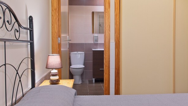 Un dormitorio con cama, una mesita de noche con lámpara, y una puerta abierta que da a un baño con inodoro y espejo.
