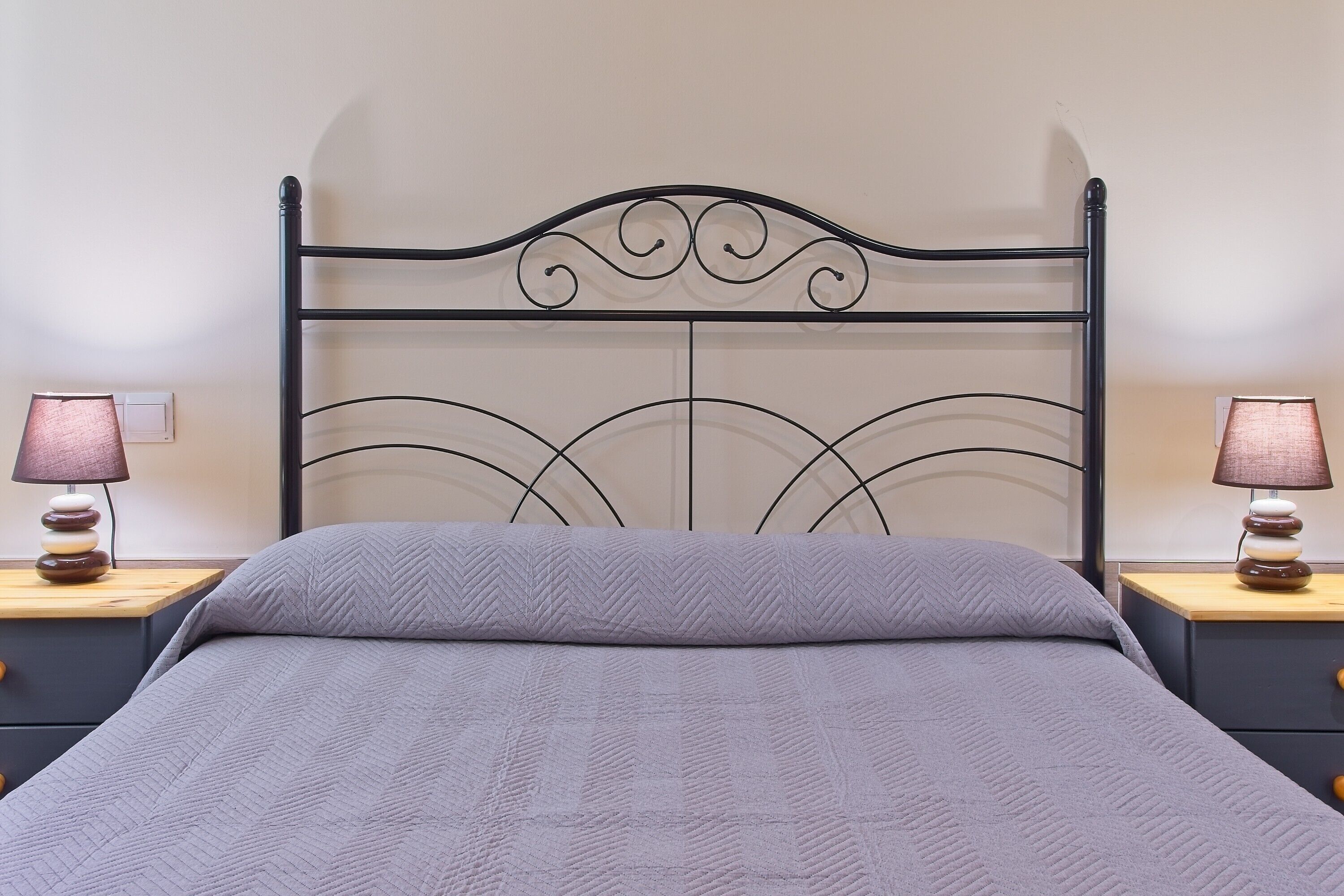 Ein Bett mit Metallkopfteil, zwei Nachttische mit Lampen und eine graue gesteppte Bettdecke.