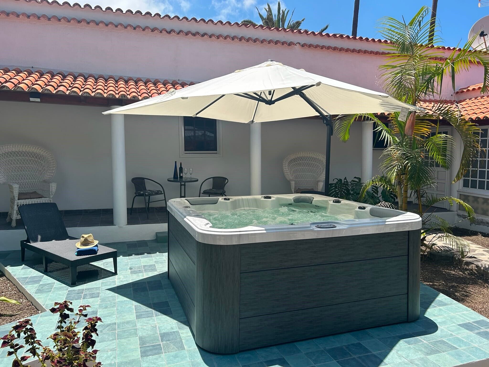 A hot tub with an umbrella, outdoor chairs, a table, and a tiled patio.