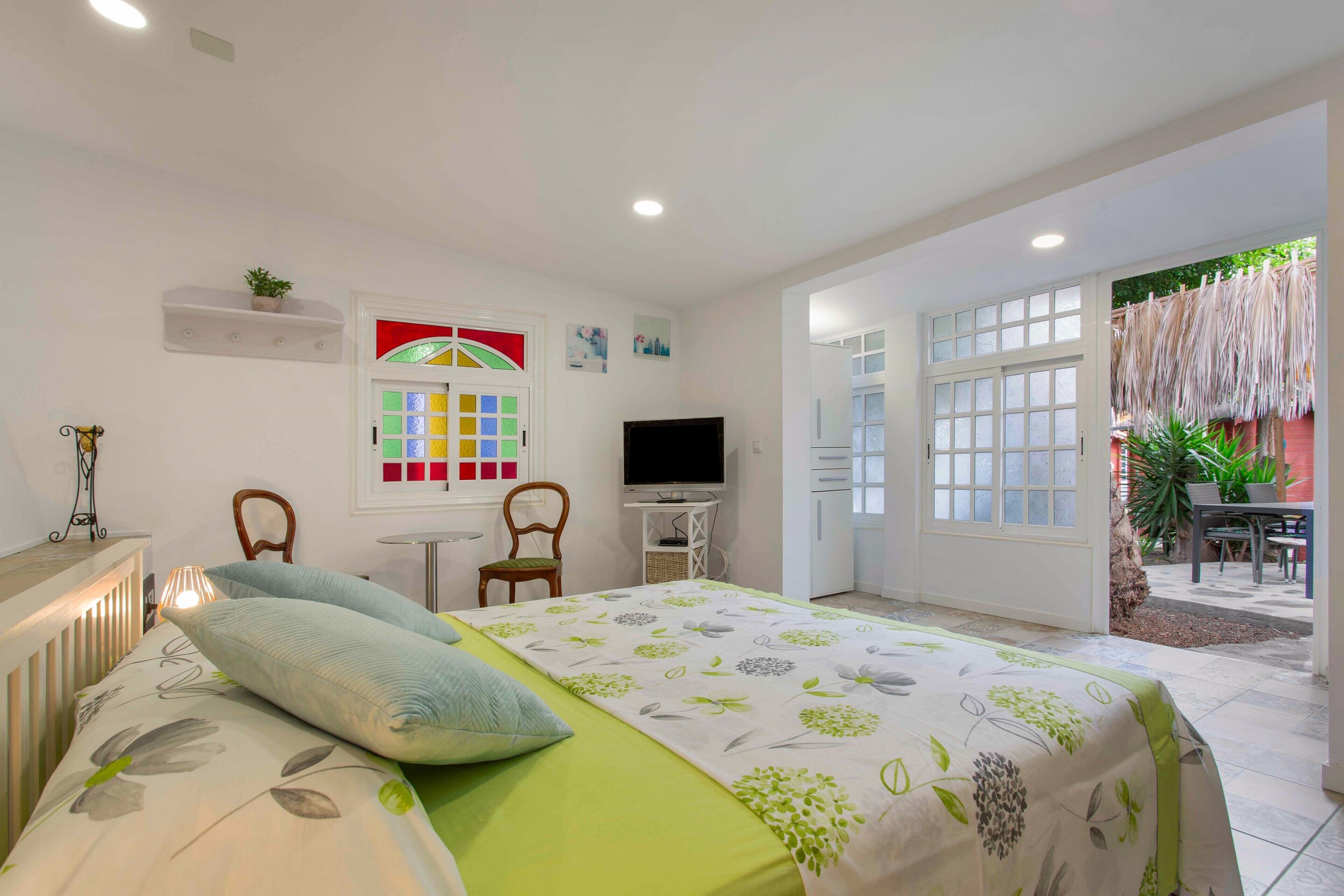 Un dormitorio con una cama, televisor, silla, mesita y ventana.