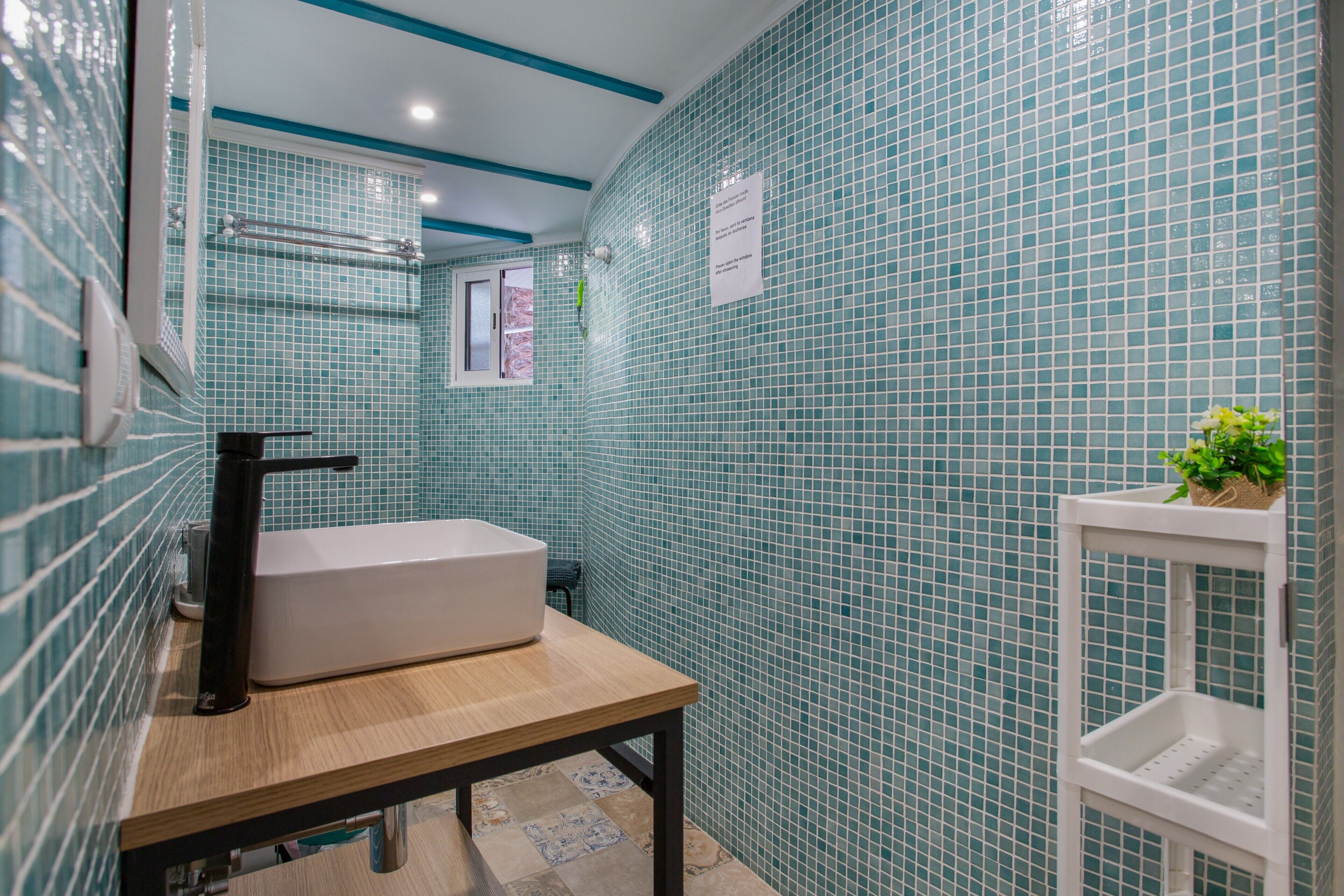 Une salle de bain avec un lavabo blanc, une table en bois, un porte-serviettes et une étagère blanche ornée d’une plante.