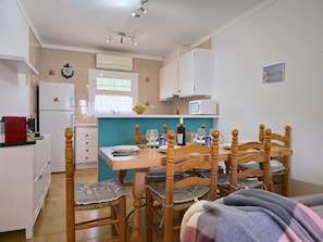 A compact kitchen with white cabinets, a refrigerator, and a microwave. There is a dining table with chairs and a window above it.