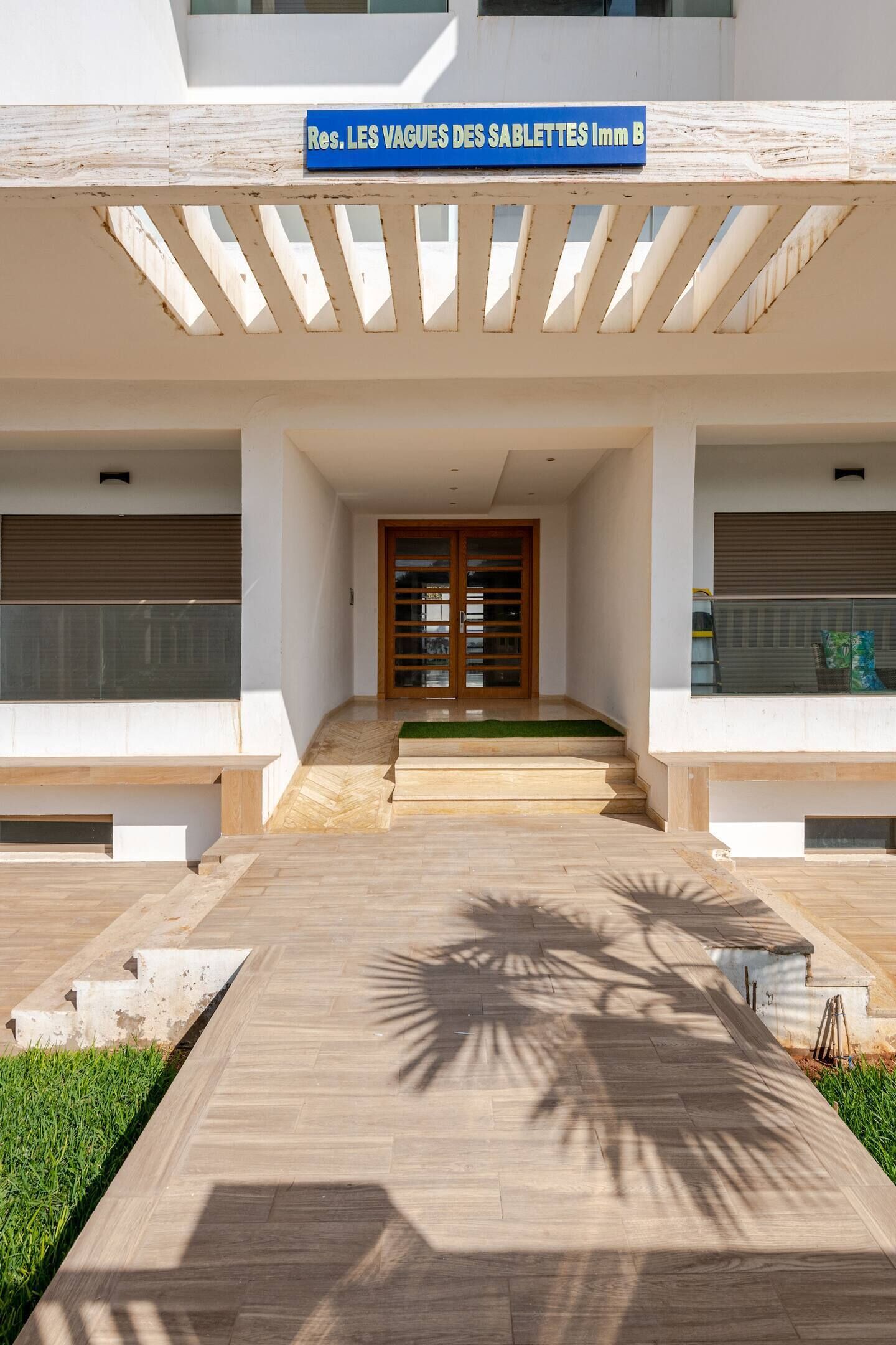 Une entrée moderne d’un immeuble, agrémentée d’une pergola en bois, d’une enseigne indiquant « Rés. LES VAGUES DES SABLETTES, Bâtiment B », et d’un chemin menant à une porte dotée de panneaux de verre.