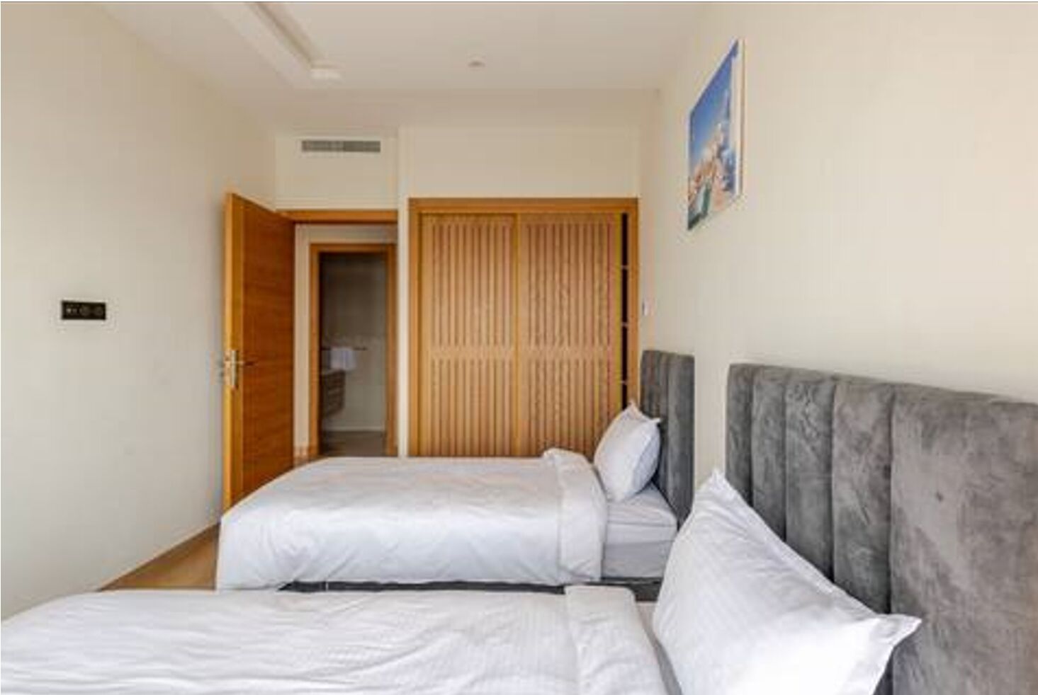 A hotel room with two beds, a wooden wardrobe, and a grey headboard.