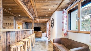 A wooden interior with a stone wall, wooden bar stools, a leather sofa, and a large window with a view of a snowy landscape. - Apartment "La Source" with Mountain Views, Private Terrace, and Wi-Fi (Valloire)