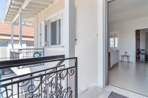Un balcón con barandilla metálica, una ventana con contraventanas y vista a un edificio con tejado de tejas rojas.
