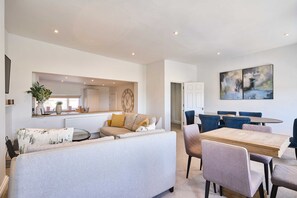 A modern living room with a sofa, dining table, and chairs. There is a kitchen area in the background with a clock and a plant. - Host Stay Albert House (Weymouth)