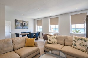 A spacious living area with a beige sofa, a glass coffee table, and a dining table with chairs. There are large windows with beige blinds, a painting on the wall, and a view of the sea. - Host Stay Albert House (Weymouth)