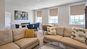 Ein geräumiger Wohnbereich mit einem beigefarbenen Sofa, einem Glastisch und einem Esstisch mit Stühlen. Große Fenster mit beigen Jalousien, ein Gemälde an der Wand und Meerblick.