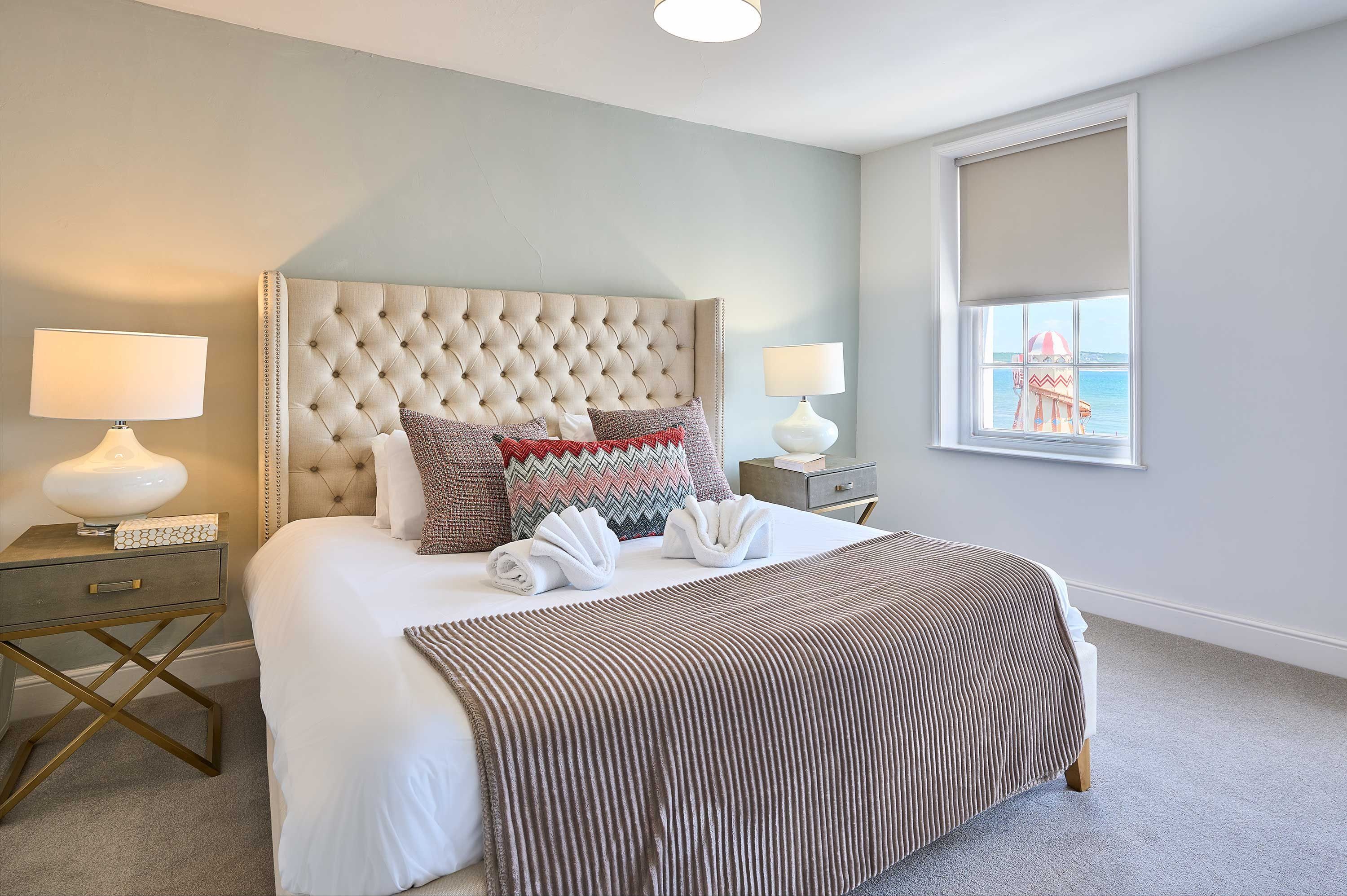A neatly made bed with a tufted headboard, two bedside tables with lamps, and a large window with a view of a building and water.