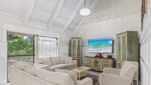 A spacious living room with a large flat-screen TV, beige sofas, a wooden coffee table, and a view of the outdoors through large windows.