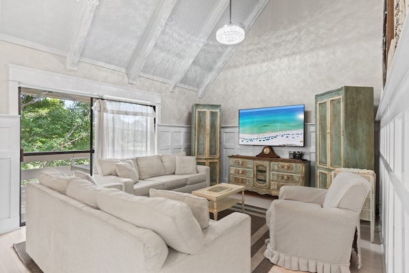 A spacious living room with a large flat-screen TV, beige sofas, a wooden coffee table, and a view of the outdoors through large windows.